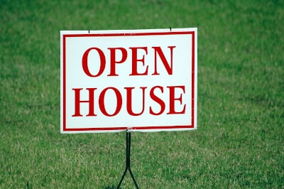A white sign with red lettering reading 'Open House' stands on a well-kept green lawn