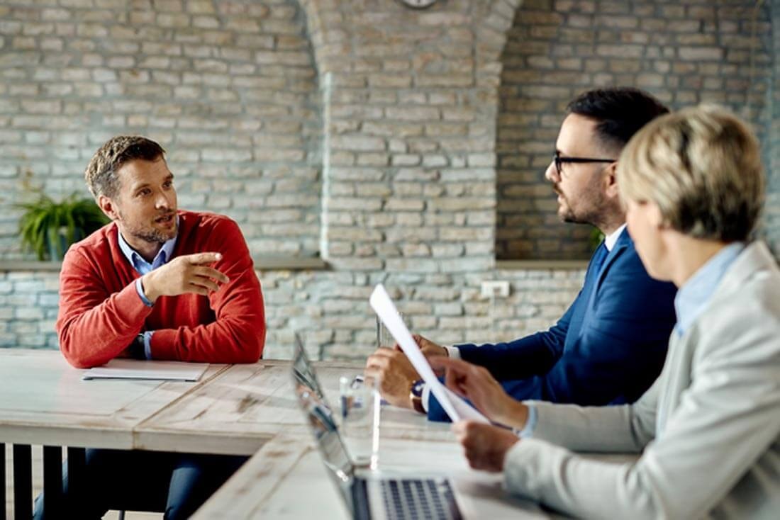 A man in a red sweater sits with a seller and an agent at a conference table explaining something, with an open laptop on the table next to the second agent