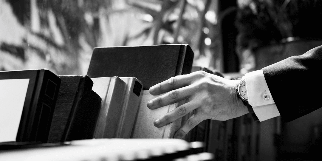 Black-and-white photograph: a man's hand in a suit with a watch reaches for a folder in a row of ring binders.