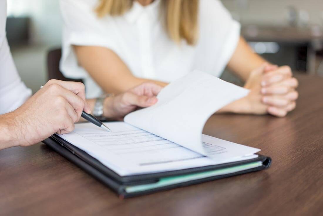 Two people sit together at a meeting table; while one listens, the other explains a detail from one of the documents lying in front of her and points to it