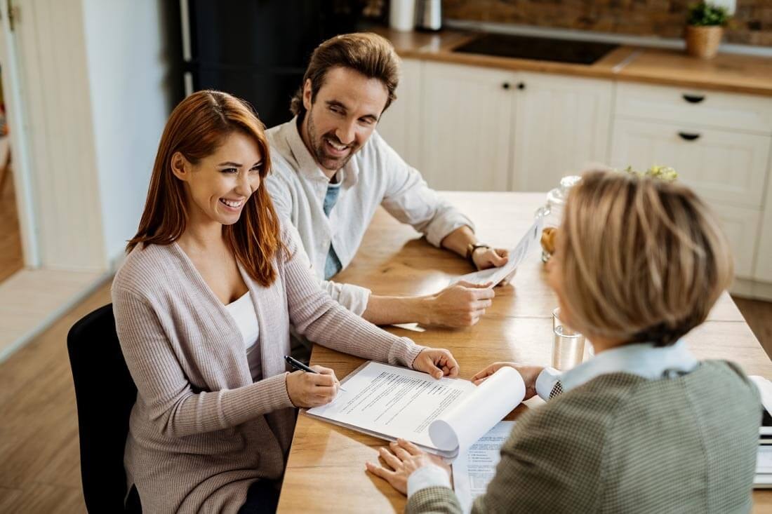 Top-down view over the shoulder of a real estate agent sitting at a kitchen table with a couple, placing a contract in front of the woman for her signature