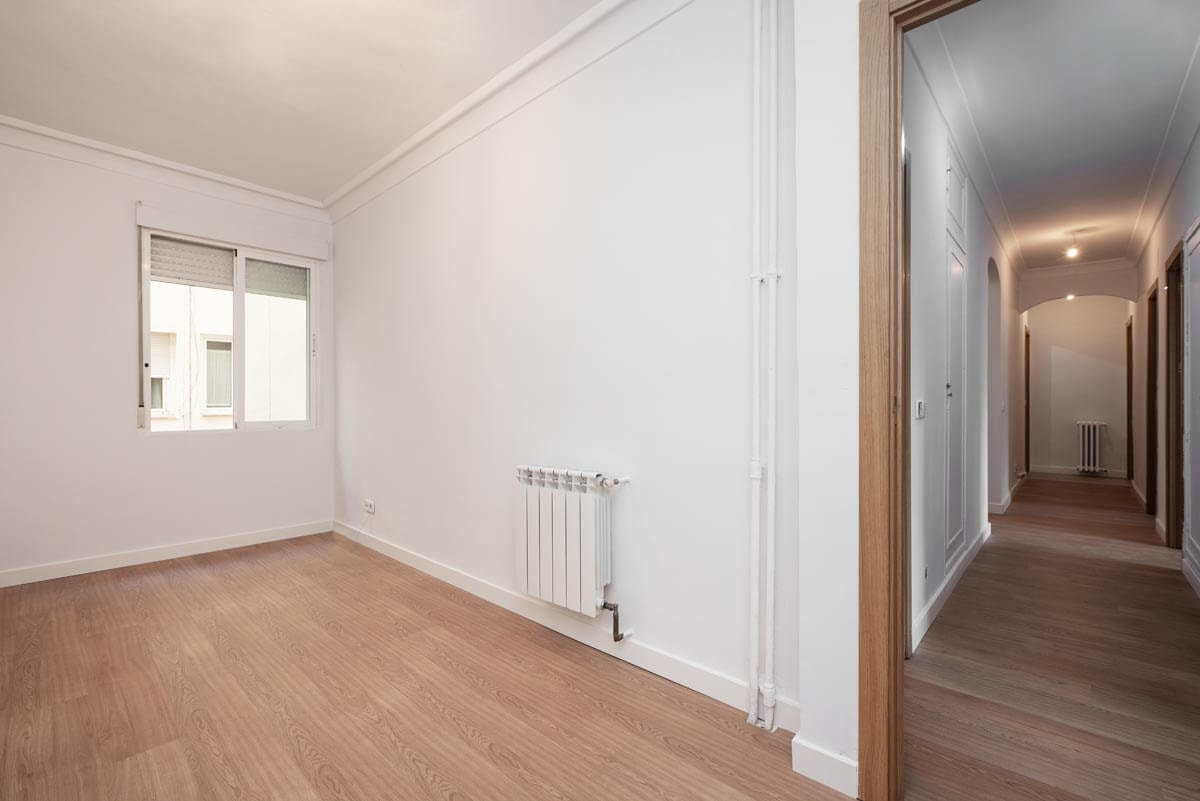 View from the corner of a room into the long hallway of an empty apartment with laminate flooring and white walls.