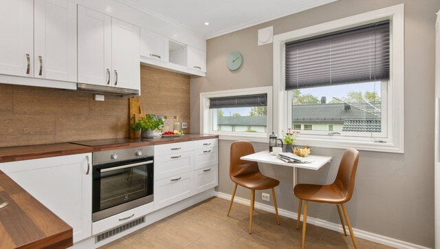 Angled view of an L-shaped kitchen unit with white cabinets and a dark worktop, a small wall-mounted table with two chairs, and two windows of different sizes on the wall.