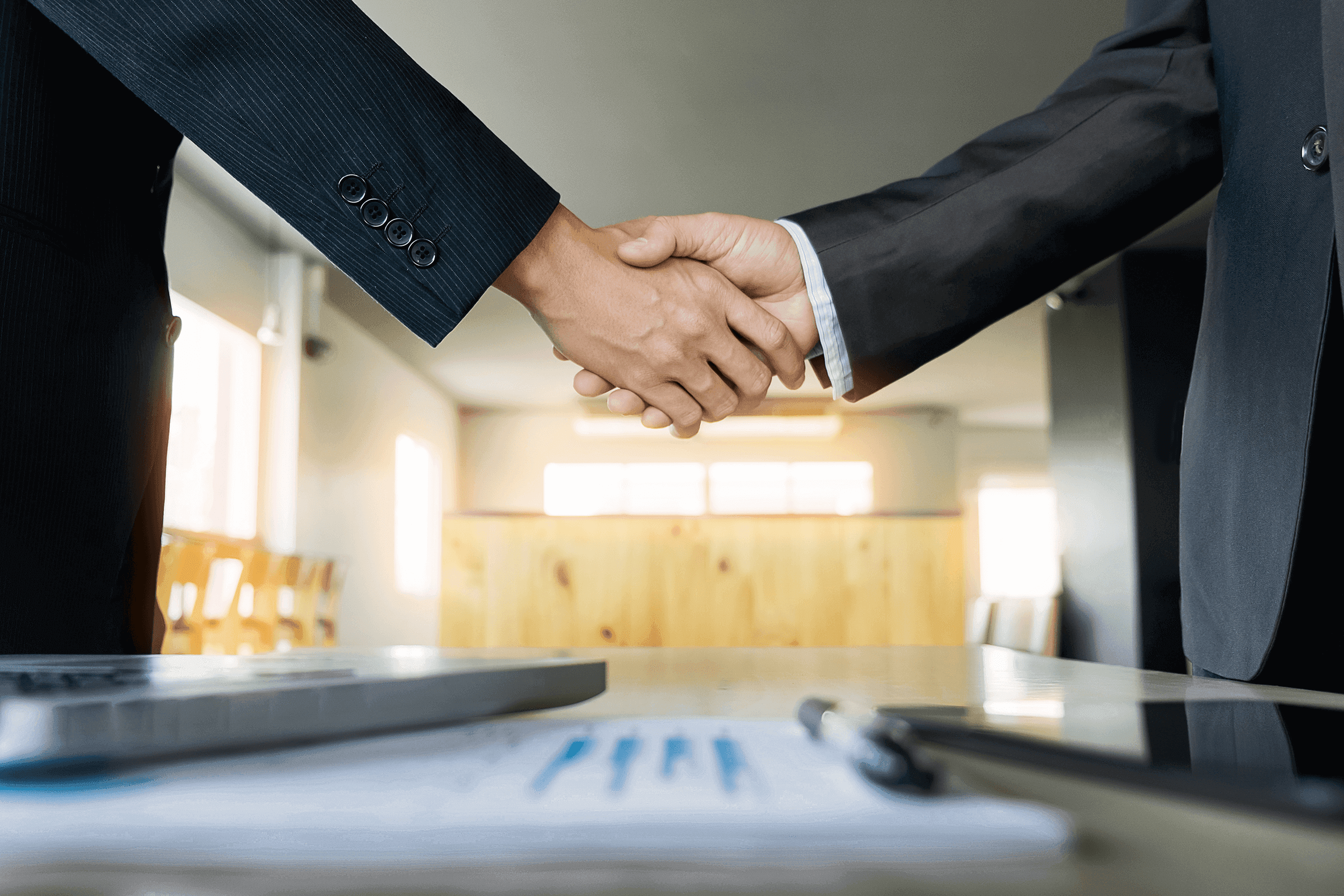 Two people in dark suits shake hands over a desk with documents and a laptop.