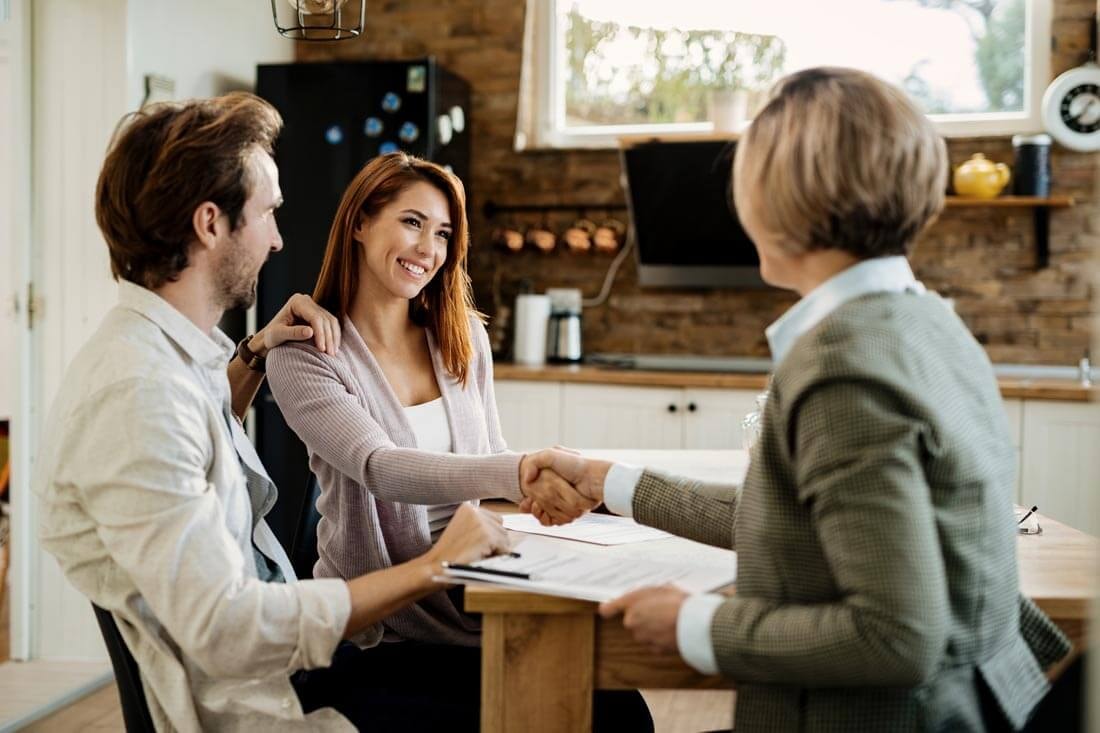 A real estate agent sits with a couple at a wooden kitchen table with several documents spread out, shaking hands with the woman