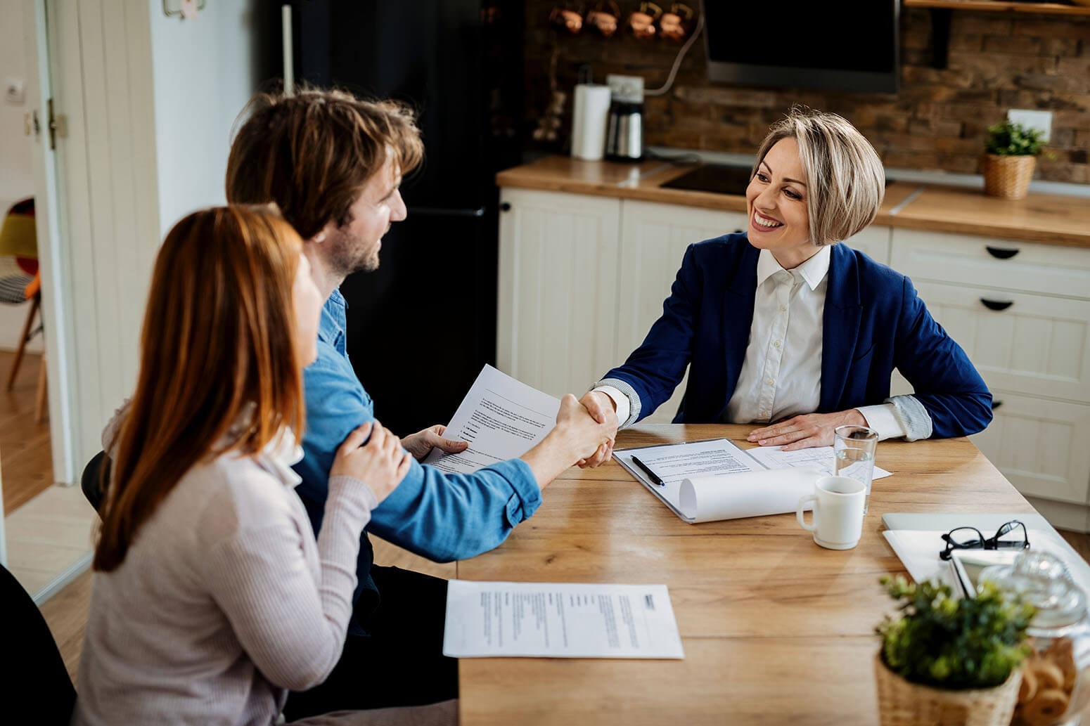 A real estate agent sits with a couple at a kitchen table with documents lying on it, shaking hands with the man