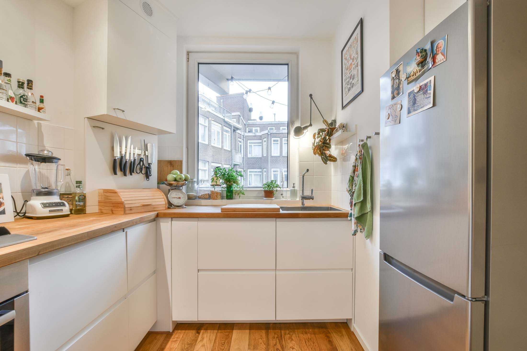 View through the doorway into a long, narrow old-building kitchen with light-coloured cabinets and a beech worktop, a large stainless steel refrigerator on the right, and the building's inner courtyard visible through the window straight ahead.