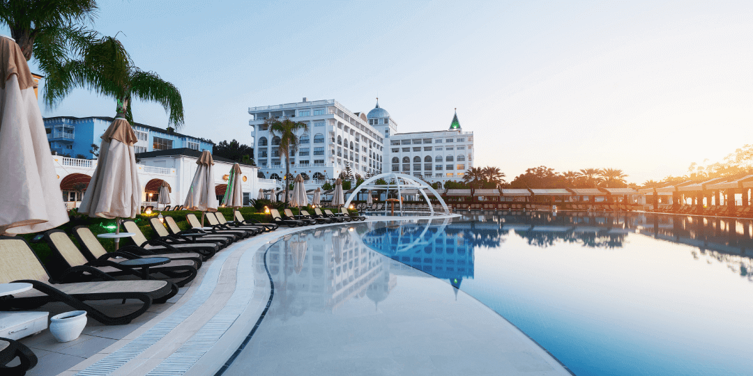 Large hotel complex with a white facade and turquoise domed tower in the early morning, with an illuminated outdoor pool, sun loungers and parasols along the poolside.