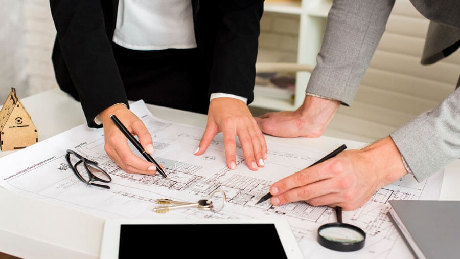 Two people standing at a table on which the floor plan of a semi-detached house is laid out, discussing details they are pointing to with pens.