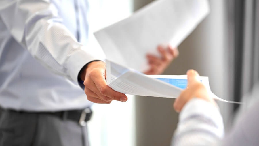 A man in a suit handing another person a document with blue print.