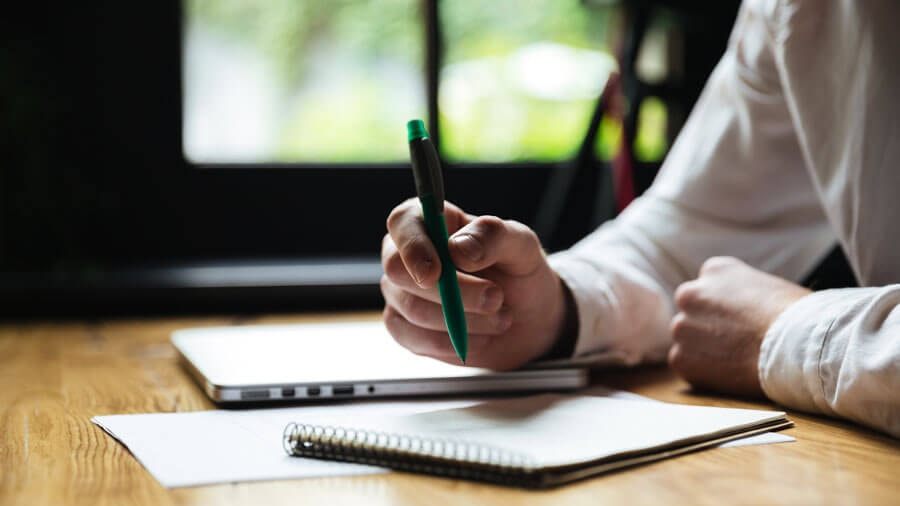 A person sitting at a wooden desk with a pen in hand in front of an open notepad, with a closed laptop beside them.