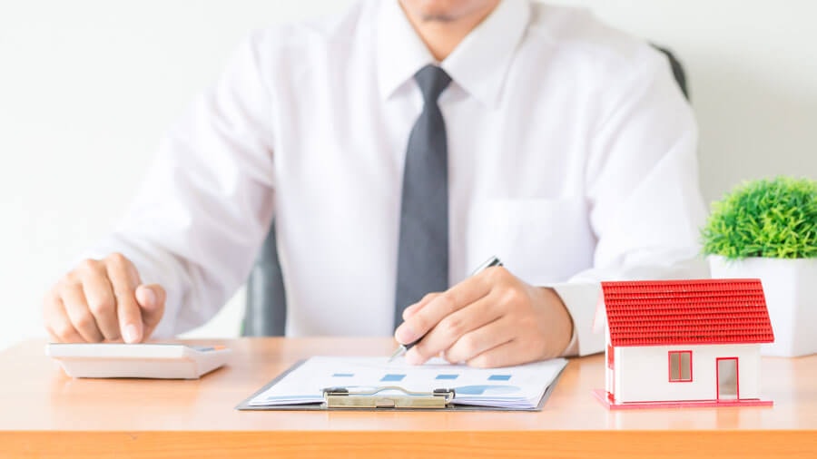 A man in a shirt and tie sitting at a desk, calculating with one hand on a calculator while noting something on paper with the other; in the foreground a miniature of a small house with a striking red pitched roof is visible.
