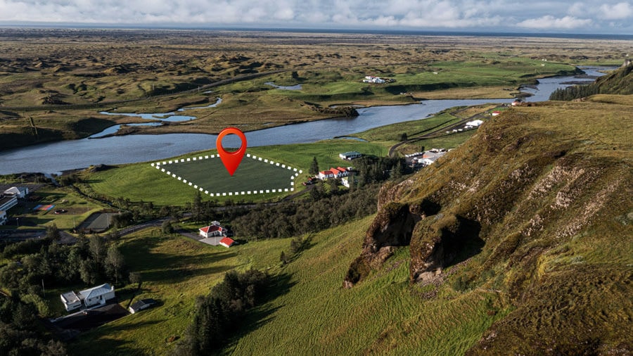Photo of a river landscape viewed from a hill, with the outline of a nearly rectangular plot of land marked schematically with a white dashed line and highlighted with a red drop marker.