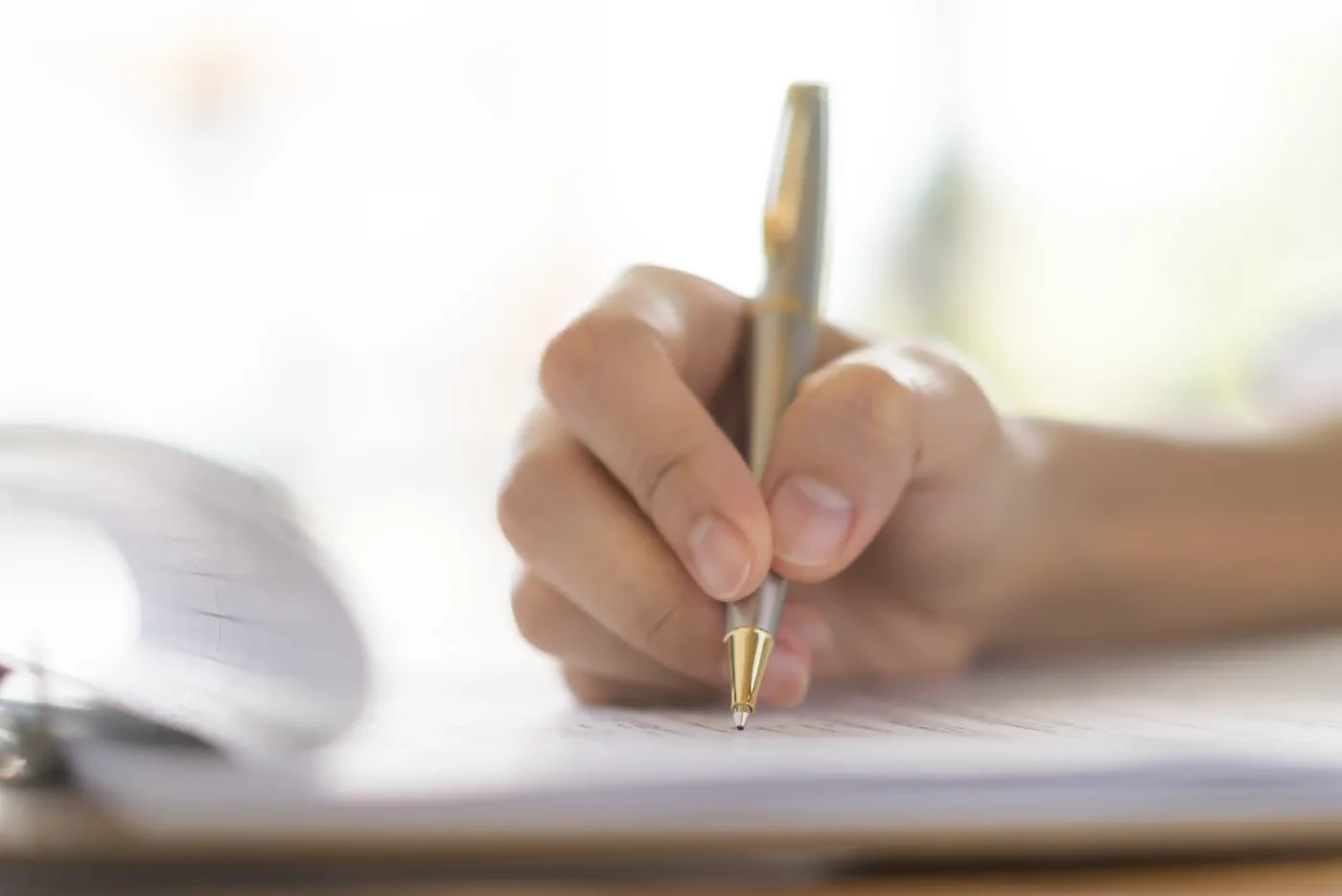 Close-up of a hand writing something with a ballpoint pen on a clipboard holding several sheets of paper.