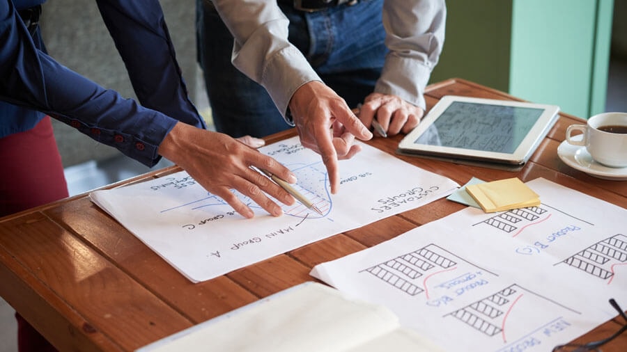 Two people leaning over a wooden table on which several diagrams and a tablet displaying a floor plan are laid out, with one hand holding a pen and pointing to the upper end of a graph curve.