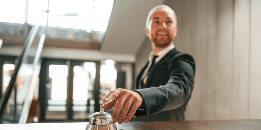 Smiling man in a dark suit ringing the reception bell of a hotel, with a bright hotel lobby in the background.