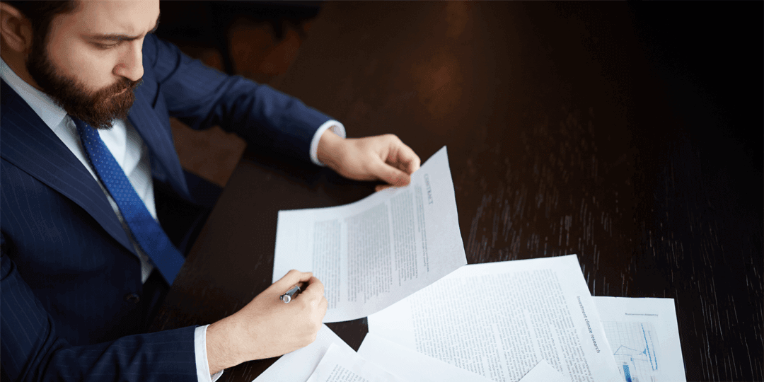 Bird's-eye view of a man in a blue suit and tie reading and carefully reviewing several documents on a dark table.