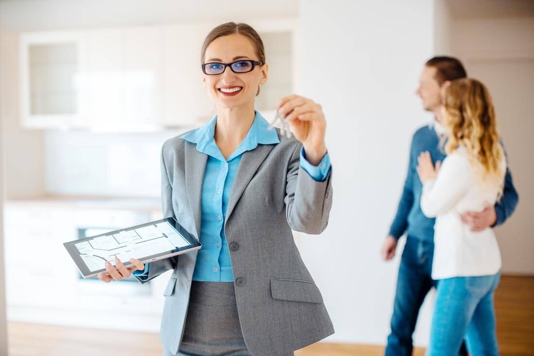A real estate agent stands in an apartment holding a tablet in her right hand and holding up an apartment key with her left, while in the background a couple stands embracing in the open kitchen, happily looking at it