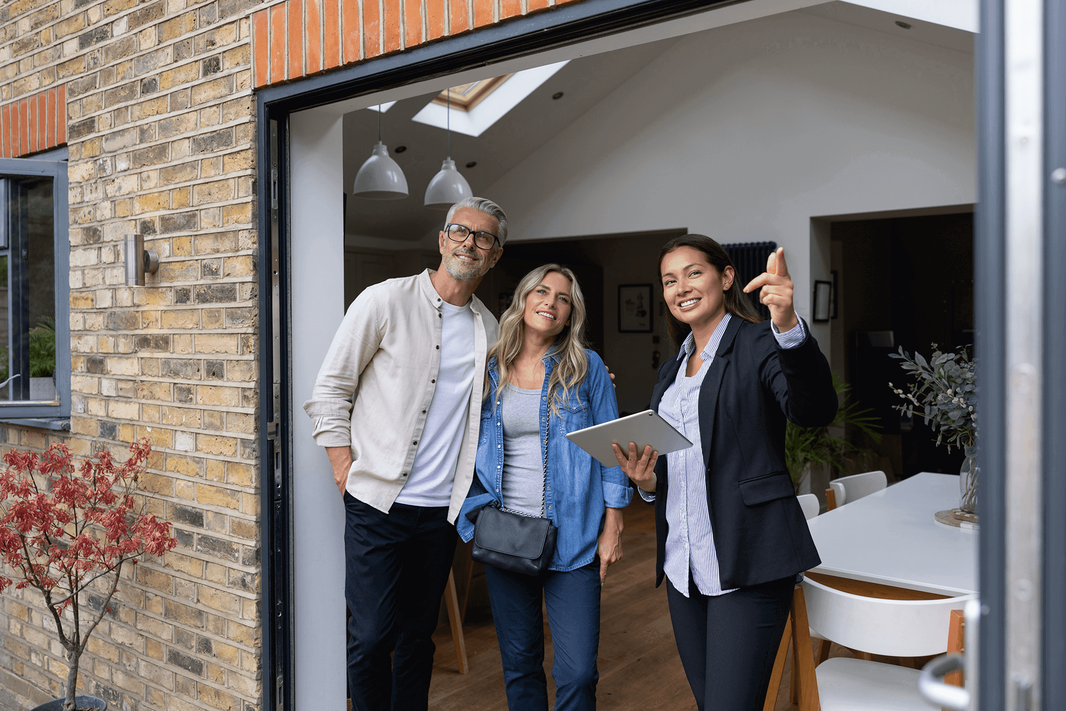 A female estate agent stands smiling with a couple in the open entrance area of a brick building and points out of the apartment; she holds a tablet in her hand.