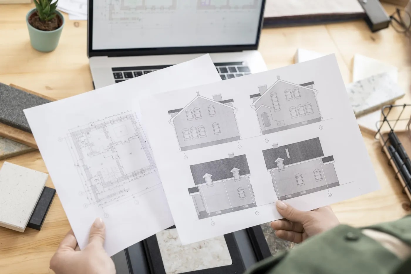 A sheet with a floor plan and a sheet with four facade elevation views of a two-story house with a gable roof, held up over a desk