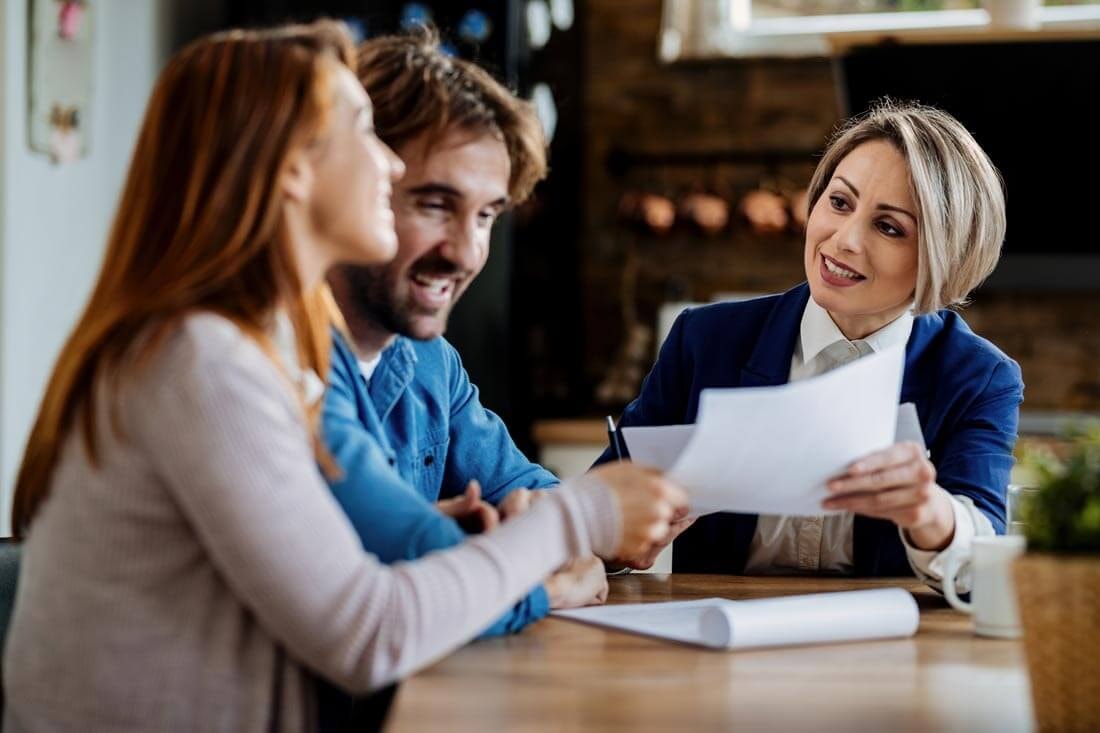 A real estate agent sits with a couple at a kitchen table, showing them some documents