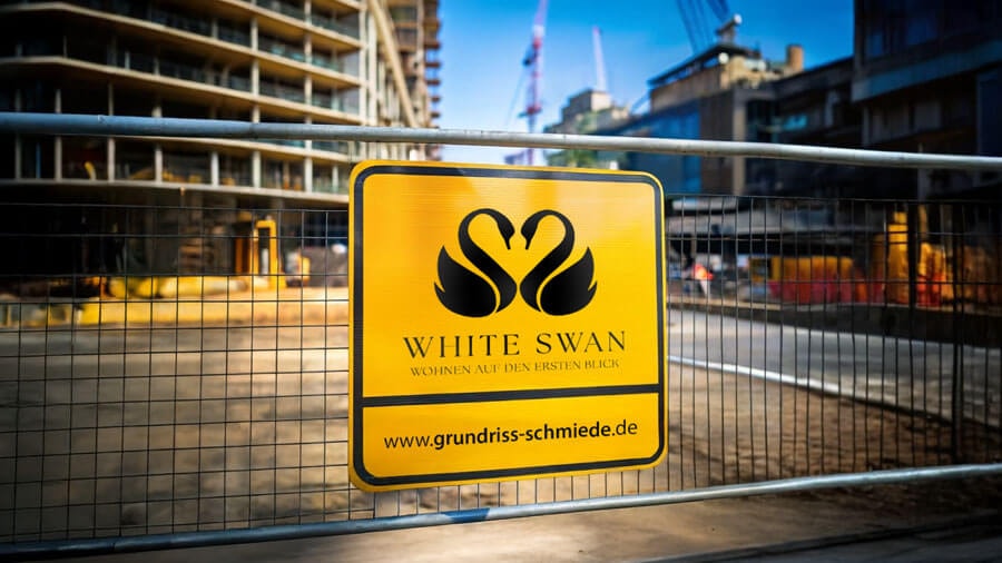 Close-up of a section of the construction fence in front of the building site, with a yellow sign showing the logo, the lettering 'White Swan', and Grundriss Schmiede's web address
