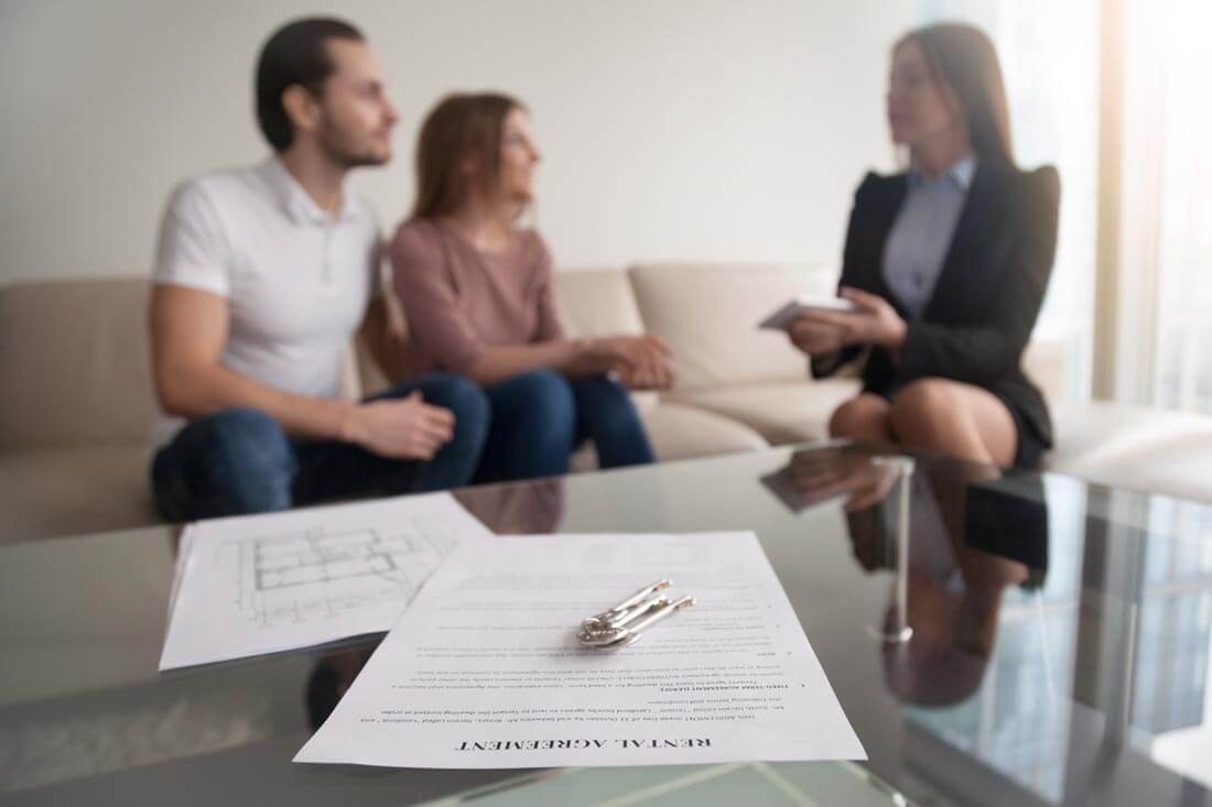 Close-up of contract documents with a key lying on top, while in the background a real estate agent sits with a couple on a light-colored couch