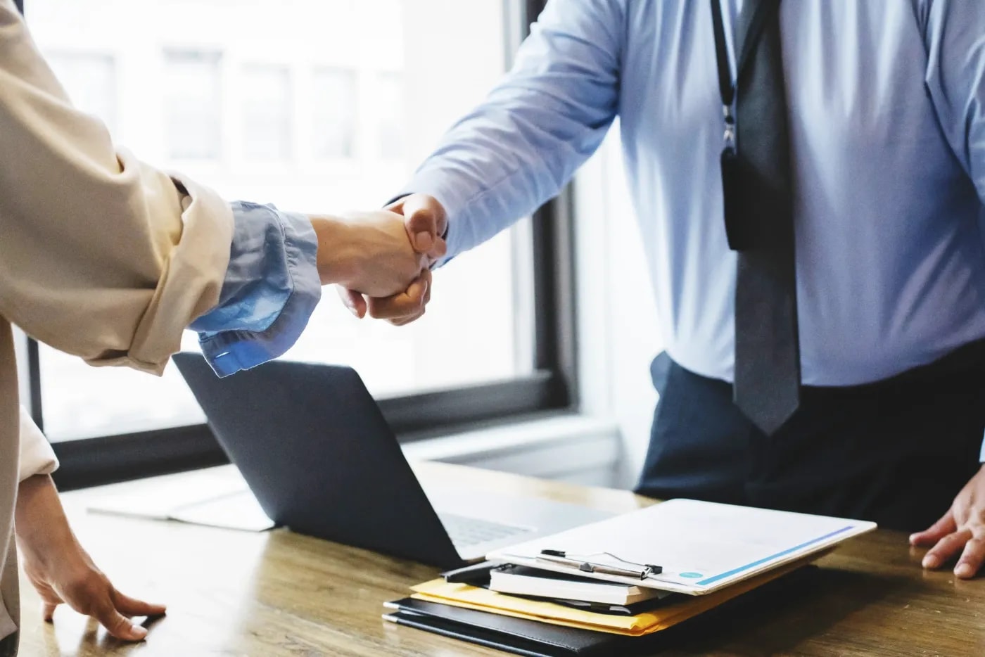 Two people standing at a desk, shaking hands across the tabletop on which some documents are stacked.