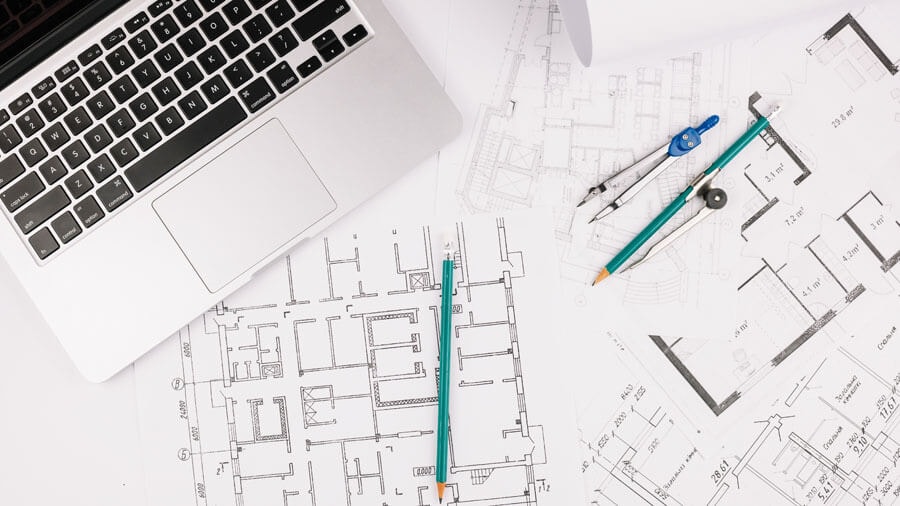 Top view of a desk with several documents alongside a laptop, with two floor plans on top, two pencils and a compass.