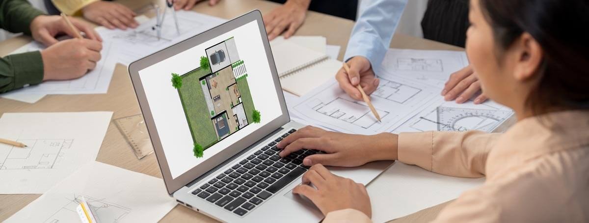 Several people stand around a square table covered with construction documents and examine them, while a person in the foreground sits at a laptop and works on a colour floor plan.