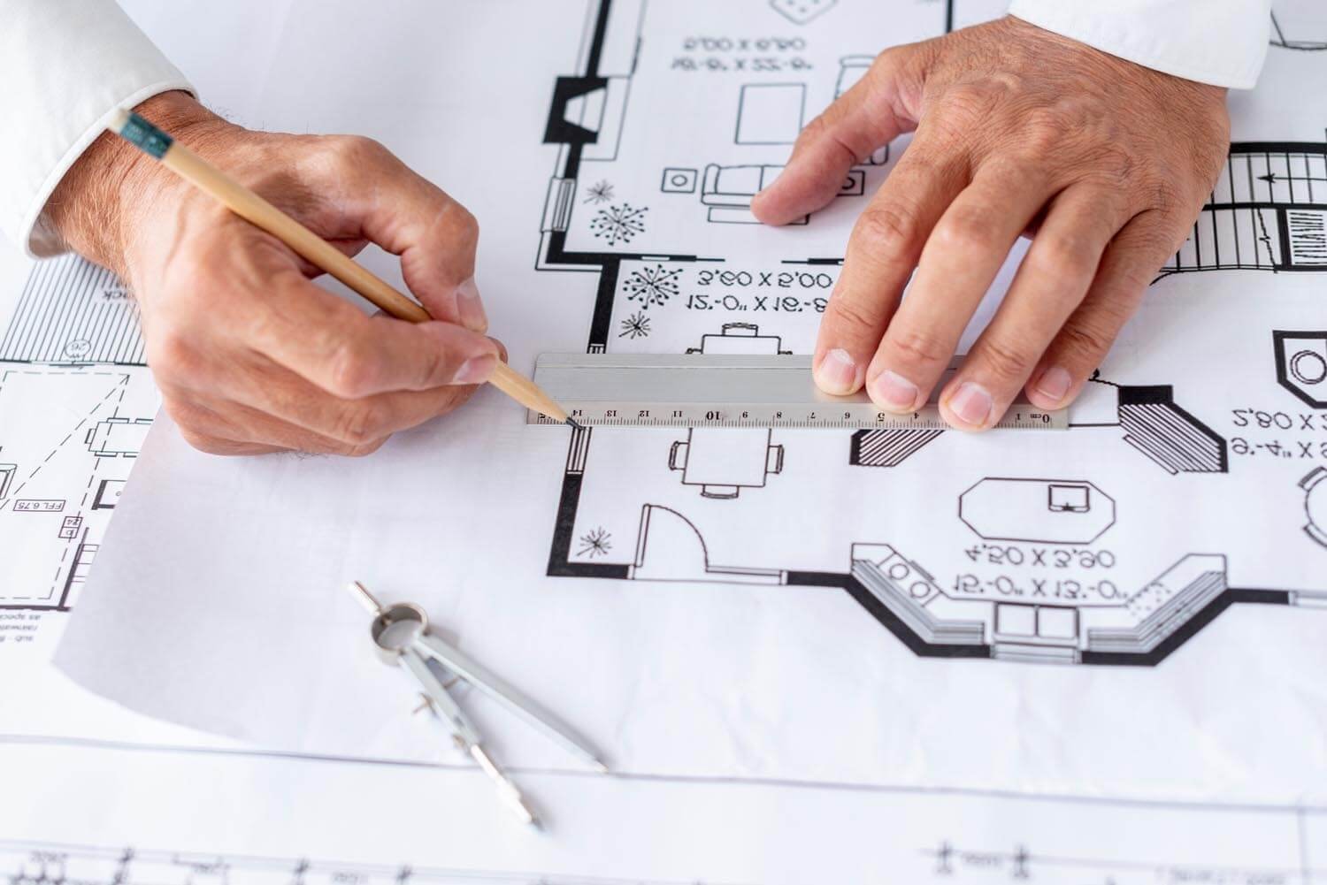 Two hands with a ruler and pencil mark a window on a floor plan at table height; a pair of compasses lies on the floor plan in the foreground