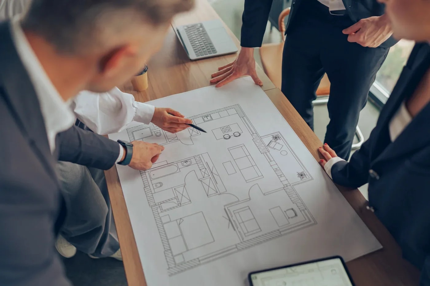 Several people stand around a wooden table, leaning over a floor plan lying on it, with one person pointing to a room with a pen.