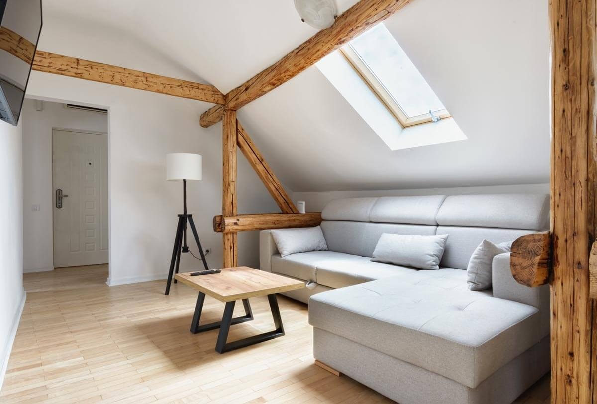 Helle Polstersitzgruppe zwischen tragenden Dachbalken aus Holz unter einer Dachschräge mit Dachfenster, gegenüber einer Wand mit TV.
