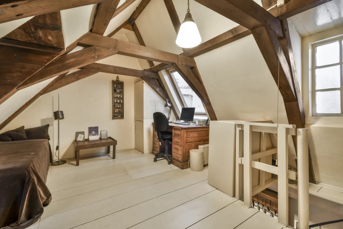 Blick aus der Raumecke in einen hohen, ausgebauten Dachstuhl mit einem Schlafbereich links und einem Arbeitsbereich mit Einbauschrank rechts vor einem Dachfenster.