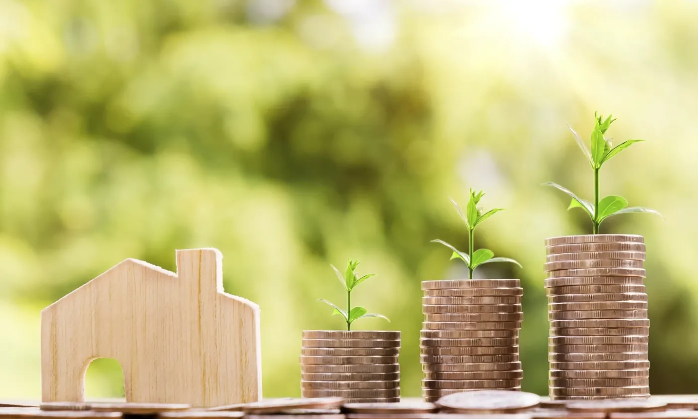 Wooden house model next to three ascending stacks of coins with seedlings, symbolising the foundation of a solid building loan