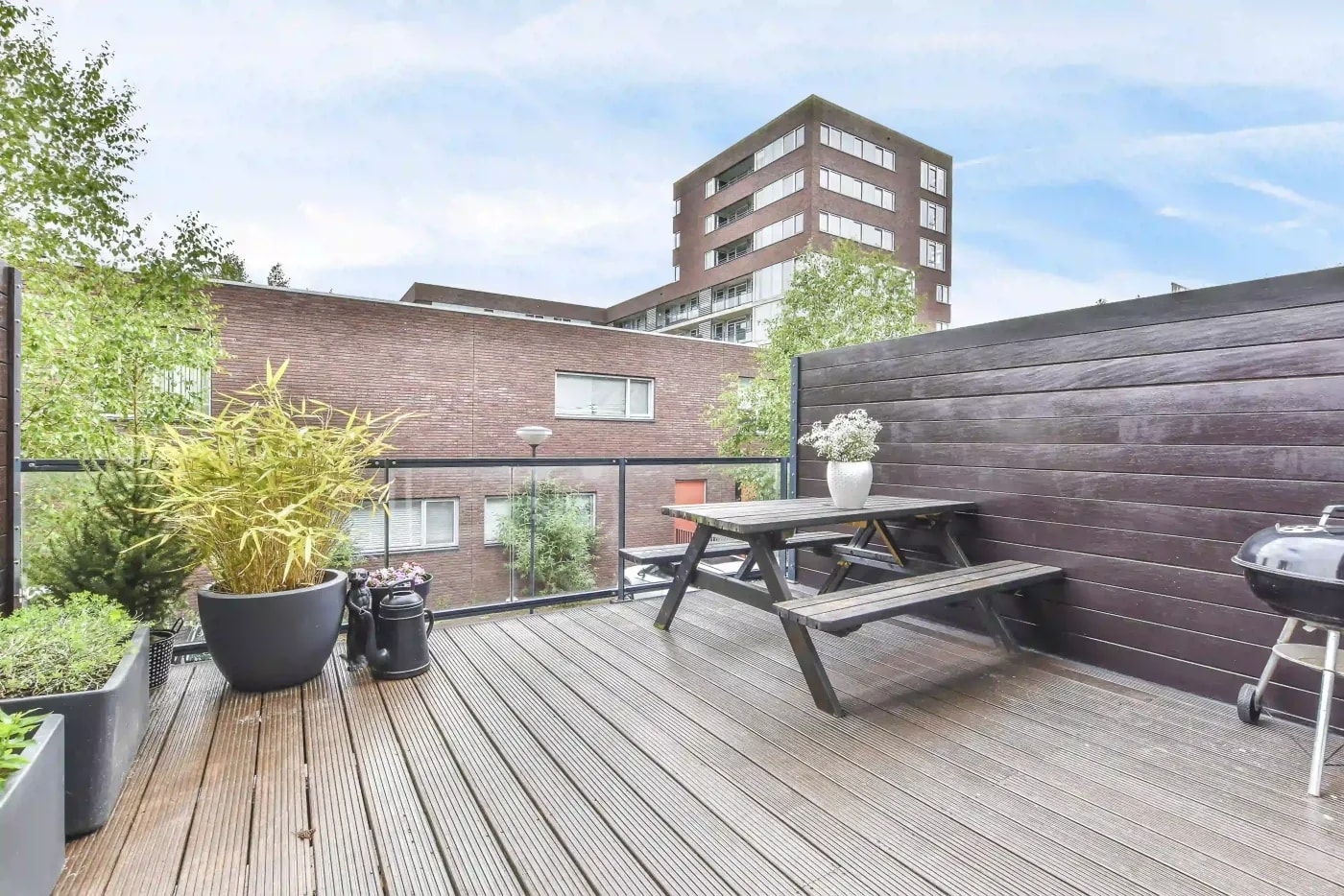View of a rooftop terrace with a wooden floor, a picnic table and grill, several green plants, a glass panel railing, and a wood panel boundary
