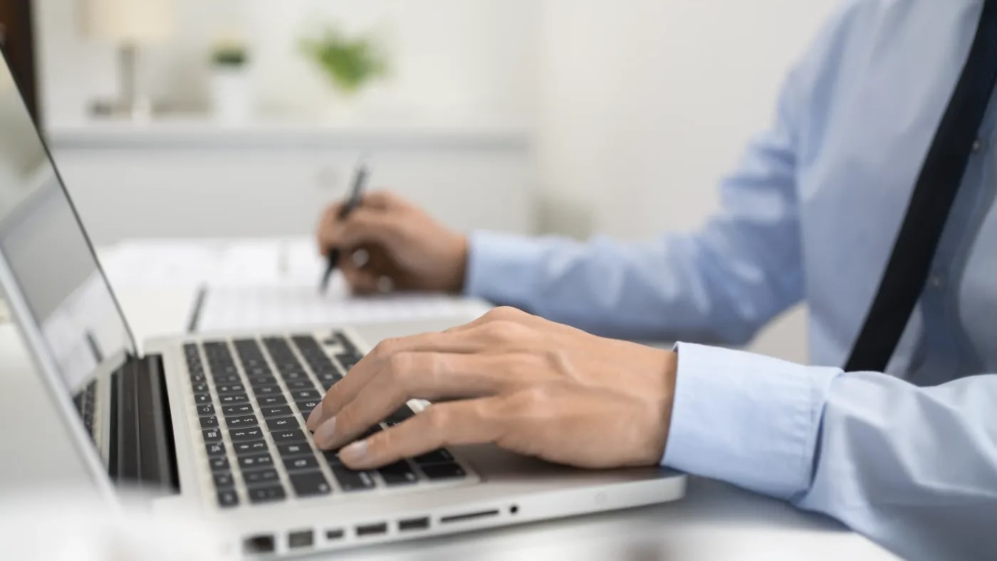 A person sitting at a desk in front of a laptop, transferring data from a sheet of paper beside them into the laptop.