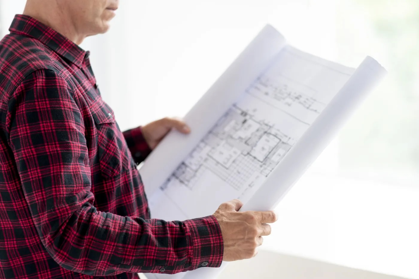 A man in a red-and-black checked shirt holds a construction drawing in front of him with both hands
