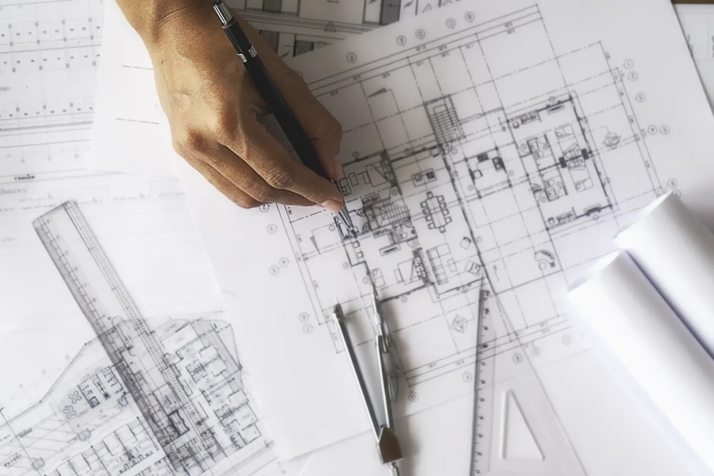 A hand with a pen draws something on a dimensioned floor plan, with rulers and a pair of compasses lying next to it