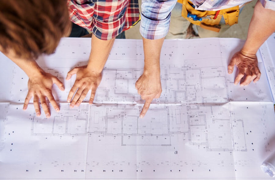 Top view: two men stand side by side, leaning over a floor plan with their hands resting on it, while one of them points to a detail