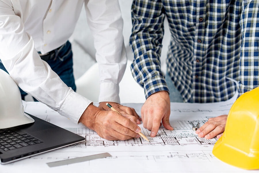 The hands of two men point to a construction drawing, with a laptop and two hard hats visible next to it