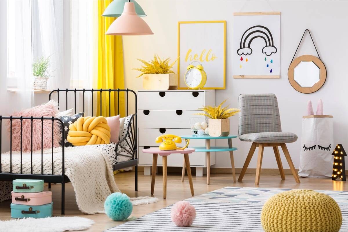 Bright children's room with a window and bed on the left, a sideboard with a chair and rainbow picture on the wall at the head of the bed, and a rug with various plush balls on the right.