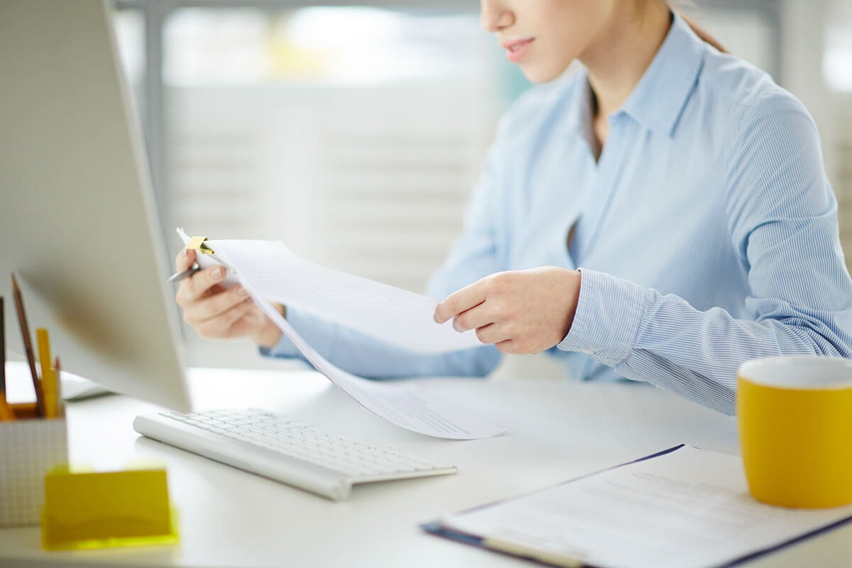 A woman sits at her desk in front of a keyboard and monitor, reviewing documents held up in her hands.