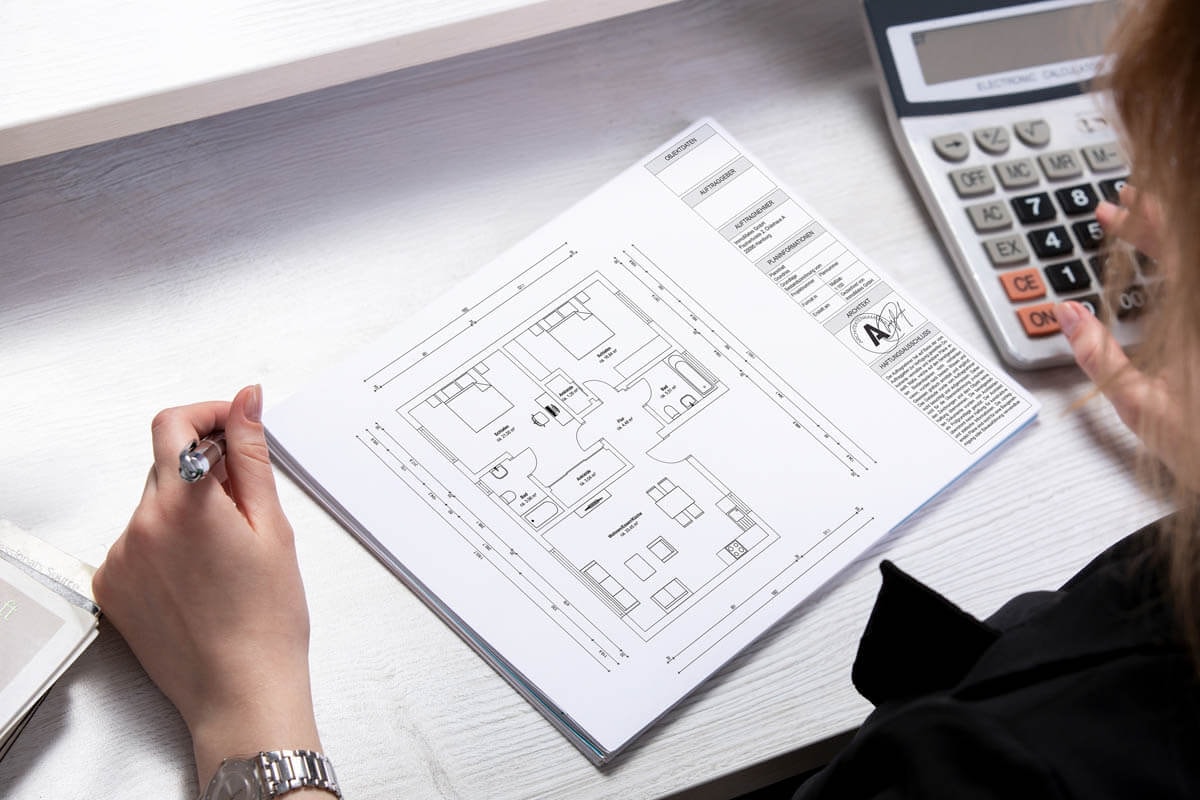 Over-the-shoulder view of a person looking at the dimensioned floor plan of a 3-room apartment in front of them, entering something into a calculator.