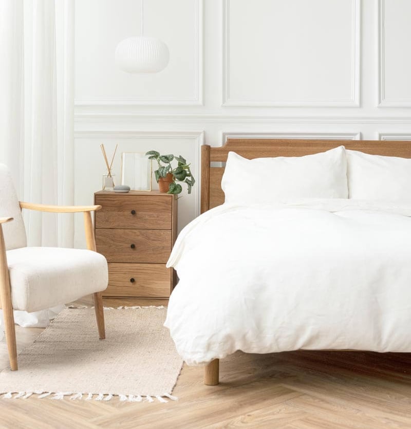 Minimalist white bedroom with a wooden bed frame, white bedding, wooden nightstand with a small plant, white upholstered armchair and decorative wall panels.