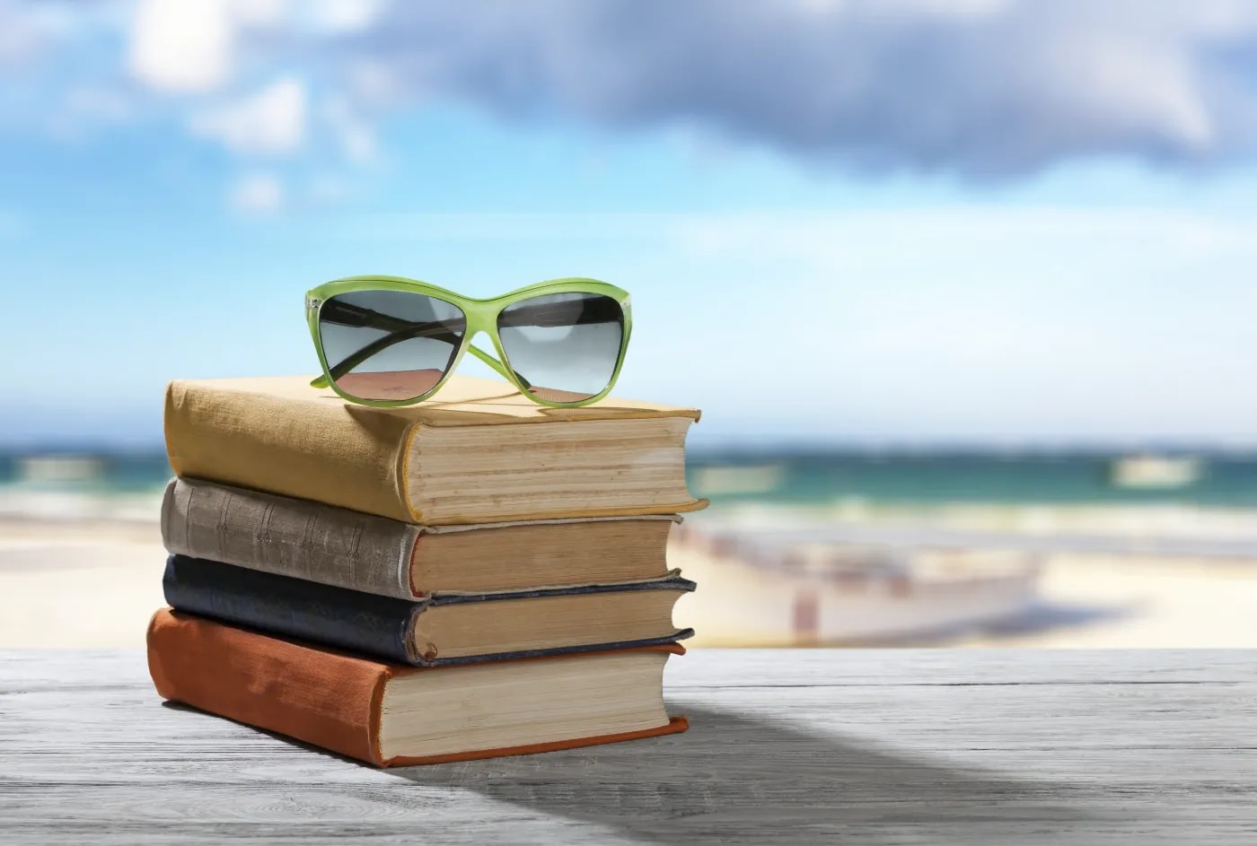 A stack of four books with a pair of sunglasses on top rests on a wooden surface in front of a blurred beach panorama in the background.