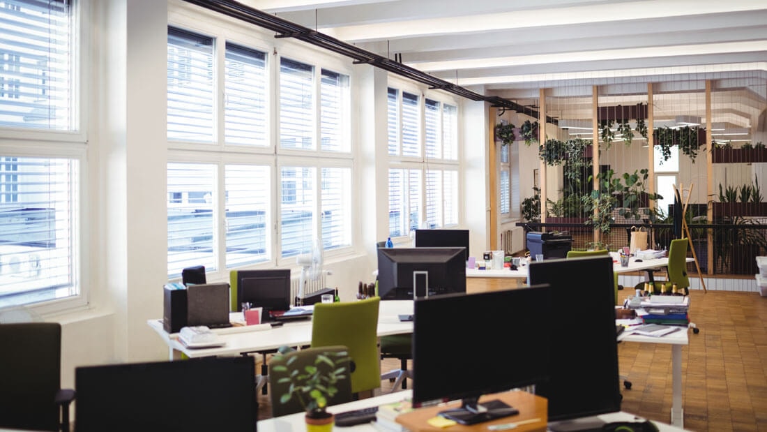Side view from the corner of an elongated open-plan office with a window front and desks arranged in groups of three, looking out onto the inner courtyard of the building complex.