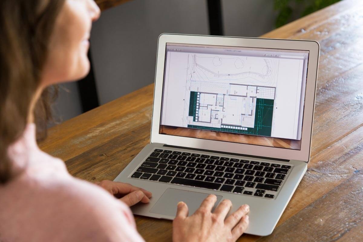 Rear view of a woman sitting at a laptop whose screen shows a building plan along with a site plan.