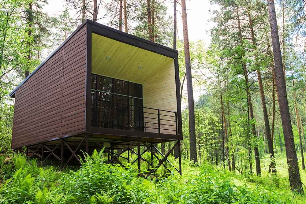 Cubist-style bungalow made of wooden slats on a steel frame perched on a densely wooded steep slope in a forest.