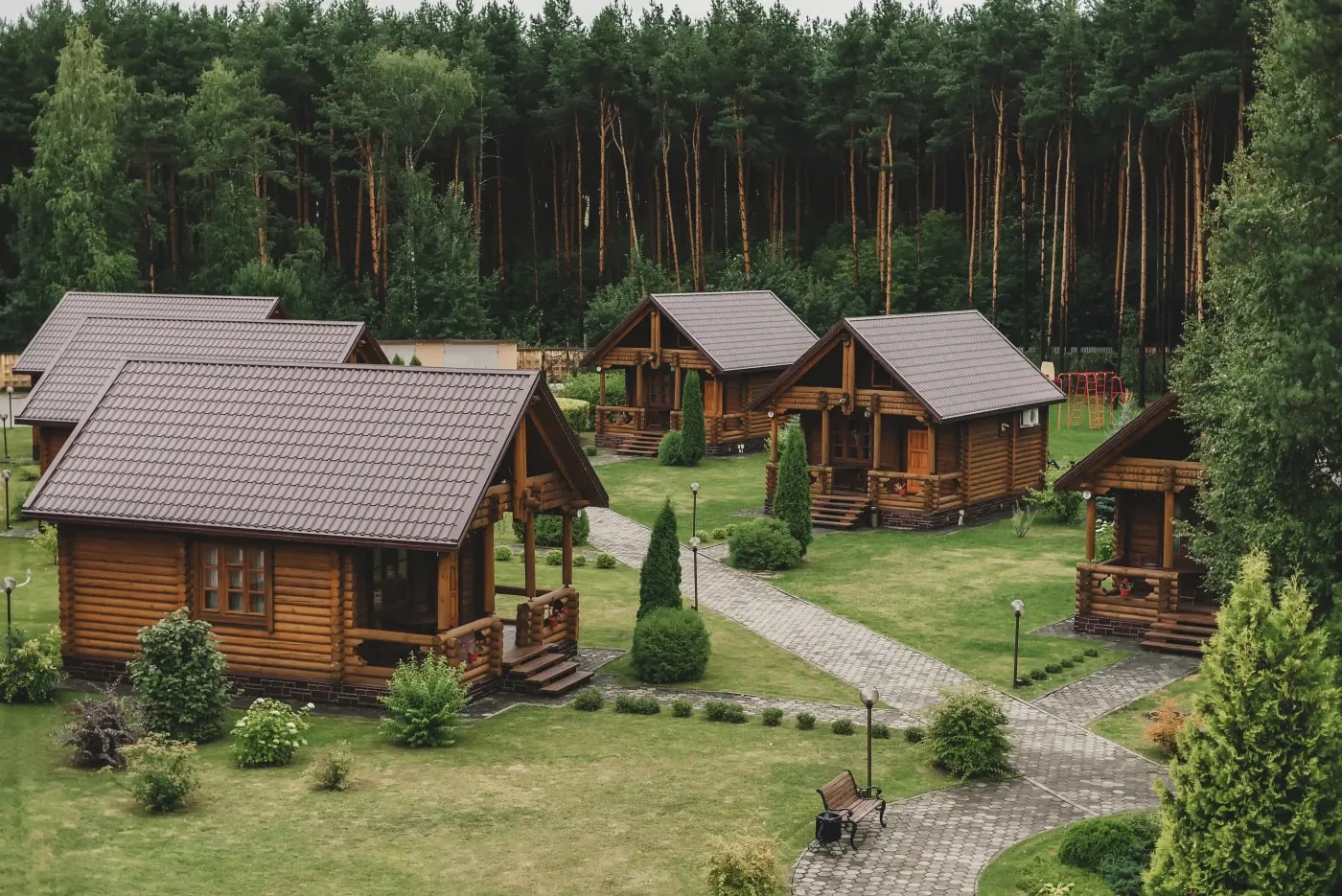 Group of identical timber bungalows with pitched roofs on a clearing in a pine forest.